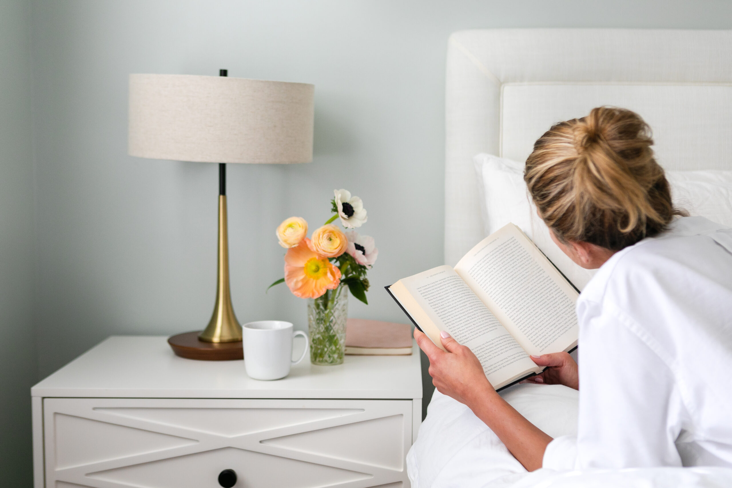 Woman over 50 lying in bed reading a book beside a nightstand with a cup of tea, fresh flowers, and a lamp—representing quiet moments with books, Audible books, or Kindle Unlimited books.