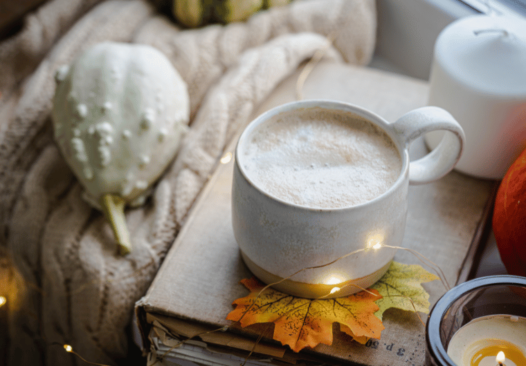 A cozy fall latte in a ceramic mug resting on a book with decorative fall leaves, knit blanket, and candles – perfect for your fall bucket list activities.