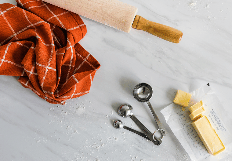 Flat lay of fall baking essentials including a red plaid kitchen towel, measuring spoons, butter, and a wooden rolling pin on a marble surface — perfect for cozy fall bucket list activities for adults.
