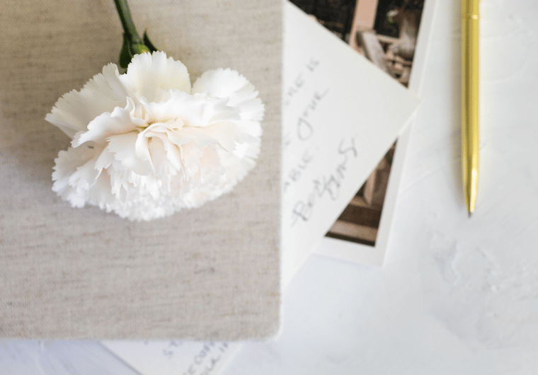 A soft white carnation rests on a linen-covered journal beside a handwritten note and gold pen on a white textured surface—capturing a calm, intentional workspace for using the Pomodoro technique.