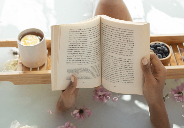 Woman relaxing in a bath, reading a book, surrounded by floating pink flowers and a cup of tea with lemon on a wooden bath tray—capturing self-care, mindfulness, and wellness for women over 50.