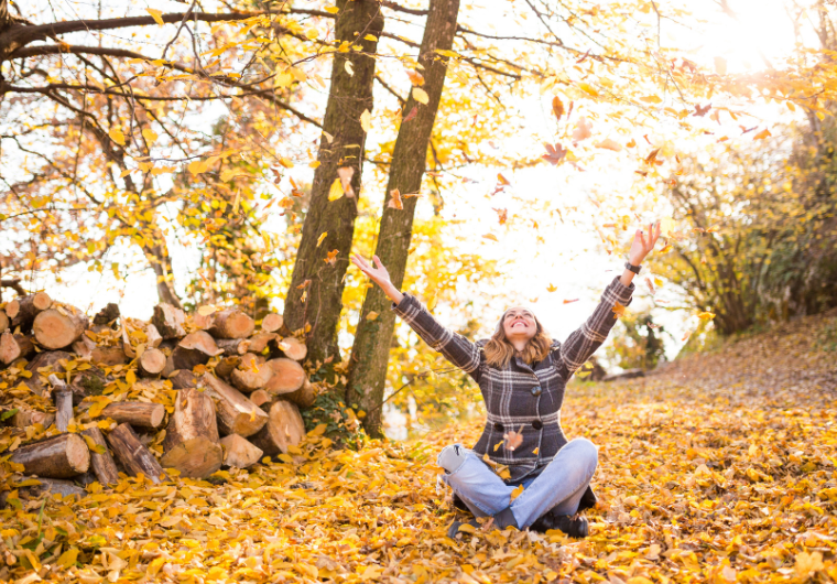Woman tossing autumn leaves joyfully in a forest, enjoying her fall bucket list moment | Angelia Mendoza