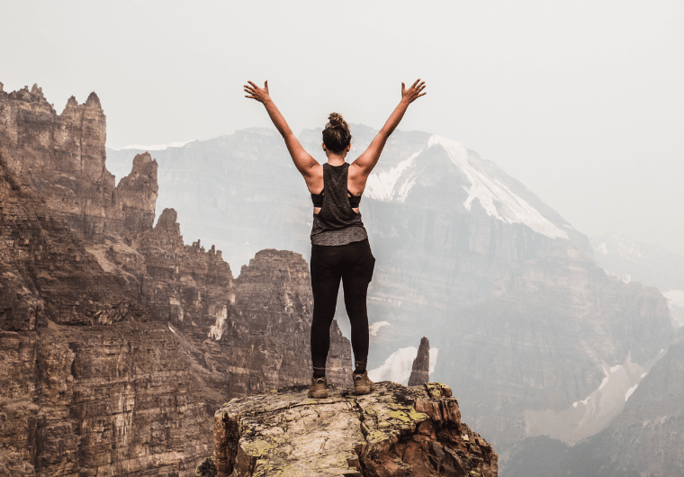 Woman standing strong on a rocky mountain peak, arms raised in triumph, overlooking rugged cliffs and snow-capped peaks in the distance. | Angelia Mendoza