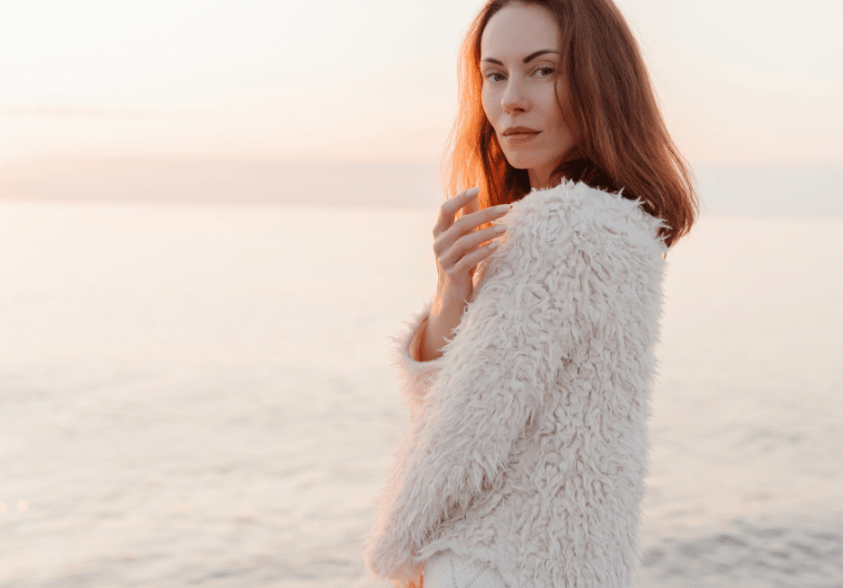 Elegant woman standing by the ocean at sunset in a soft, textured sweater, radiating confidence and beauty as she embraces how to age gracefully.