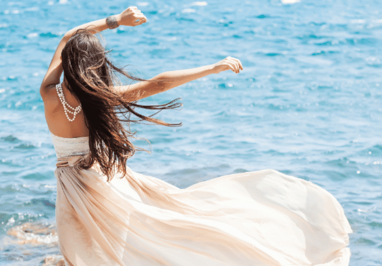 Woman in a flowing dress with long hair and pearl jewelry, standing by the ocean, arms raised in freedom and joy.
