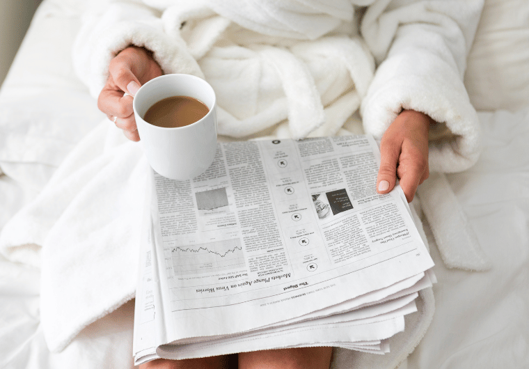 Person in a white robe sitting on a bed with coffee and a newspaper, reflecting on life and realizing it’s okay to be the villain in someone else’s story.