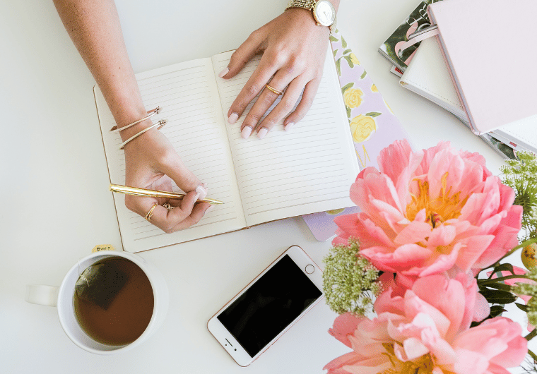 Woman writing in a journal at a bright, feminine desk with coffee, flowers, and notebooks—an inspiring scene for mid-year journal prompts for women over 50.