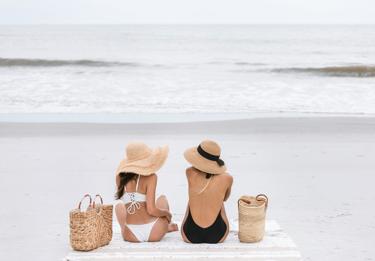 Two women in sun hats relaxing on a beach blanket by the ocean, enjoying a calm day with beach bags beside them—ideal for a relaxing getaway with essentials like a portable blender for healthy drinks.