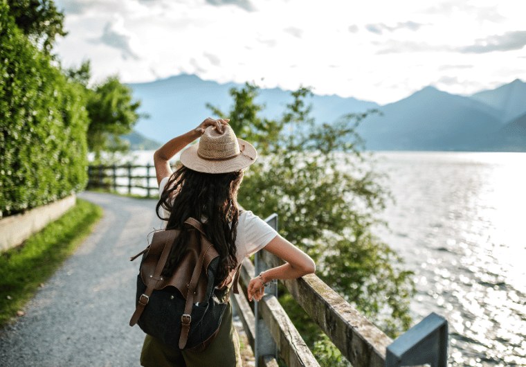 Woman traveling with backpack and sunhat walking by lakeside trail with mountain views, representing portable blender for travel lifestyle. | Angelia Mendoza