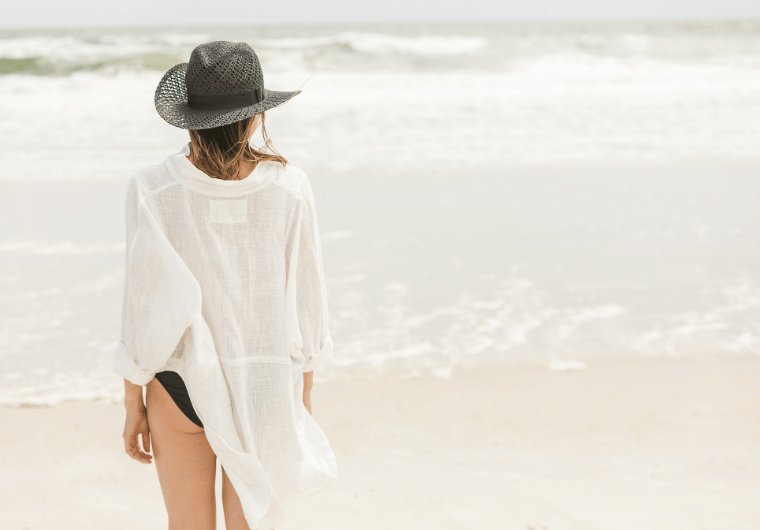 Woman in a white cover-up and black sunhat standing alone on a peaceful beach, symbolizing healing and the journey to recover after emotional abuse