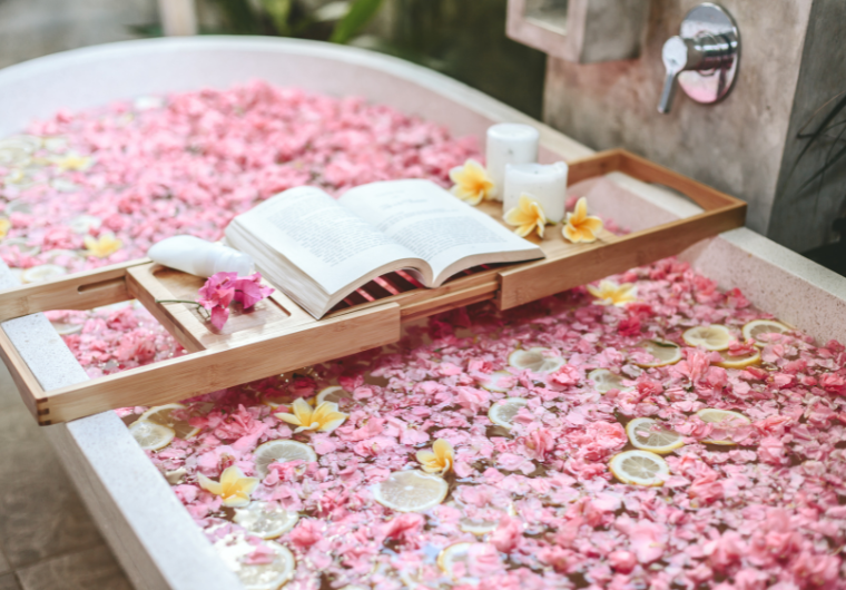 A relaxing floral bath filled with pink petals, floating lemon slices, and an open book on a wooden bath tray with candles and flowers. | Angelia Mendoza | self-care essentials for women