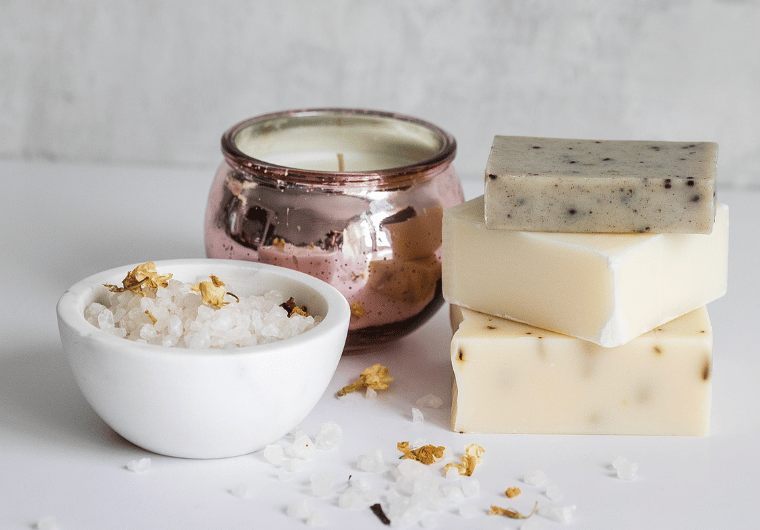 Flat lay of self-care essentials including natural soap bars, bath salts, and a rose gold candle, perfect for creating a relaxing home spa experience.