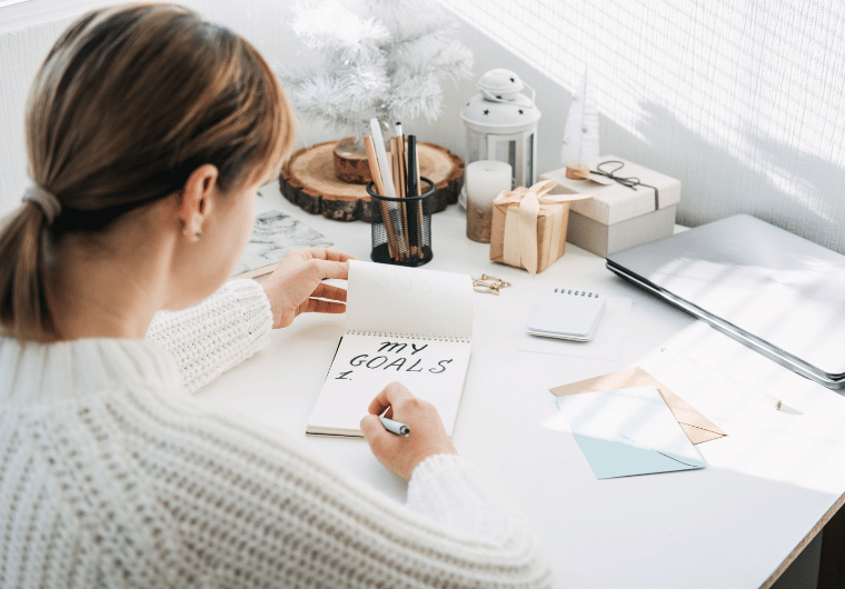 Woman in a white sweater writing “My Goals” in a notebook at a sunlit desk decorated with candles, gifts, and stationery.