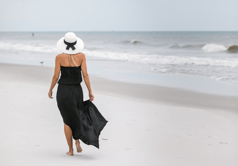 Woman in a flowing black dress and white sunhat walking along the beach, symbolizing freedom and renewal, with the message “never too late to start over.”
