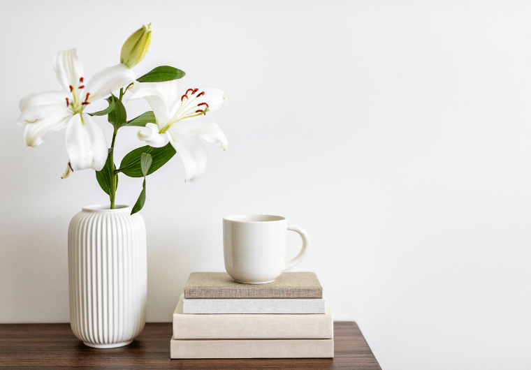 A calming flat lay of a white vase with lilies, a neutral coffee mug, and stacked books on a wooden table, symbolizing self-help books for women focused on rebuilding confidence and empowerment.